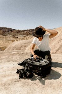A man arranges camera equipment in a desert setting, wearing a hat and sunglasses.